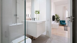 Ein modernes Badezimmer mit Toilette, Waschbecken und Spiegel. Im angrenzenden Wohnbereich befindet sich eine Pflanze und ein Türkis-farbener Sessel.