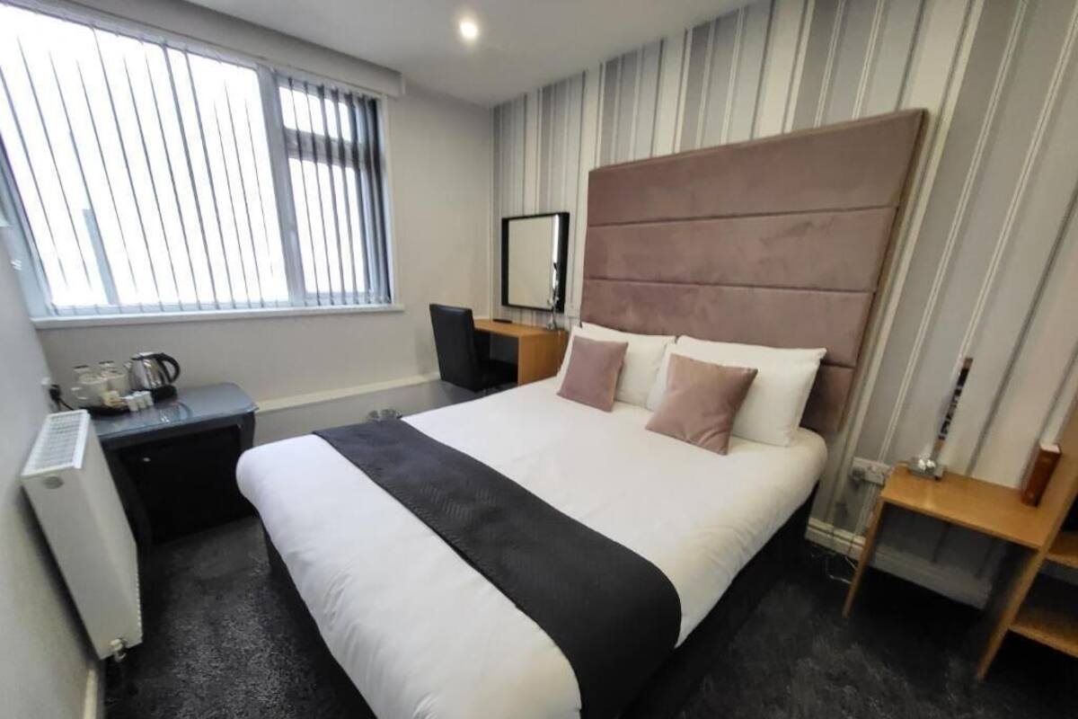 Une chambre d’hôtel avec un grand lit, un bureau, une chaise, un radiateur et une fenêtre avec des stores.