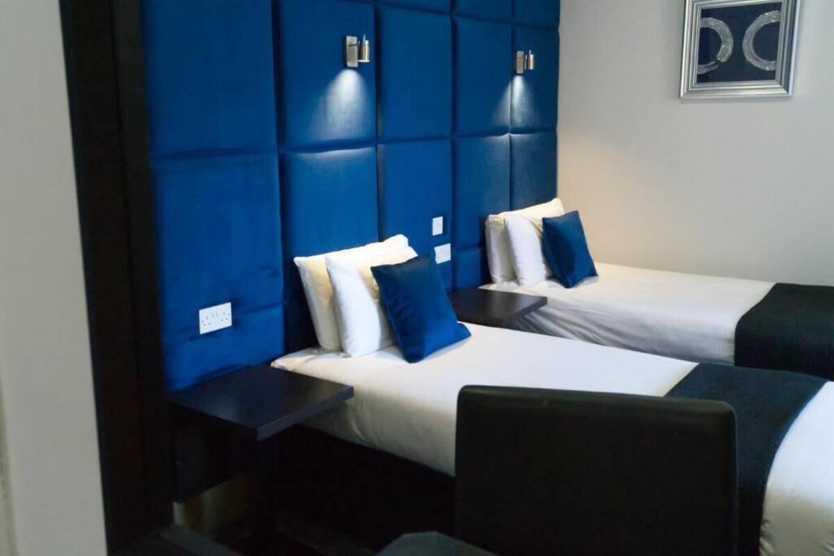 Une chambre d’hôtel avec deux lits, chacun agrémenté de draps blancs et d’oreillers bleus. Des appliques murales sont fixées au-dessus des lits.