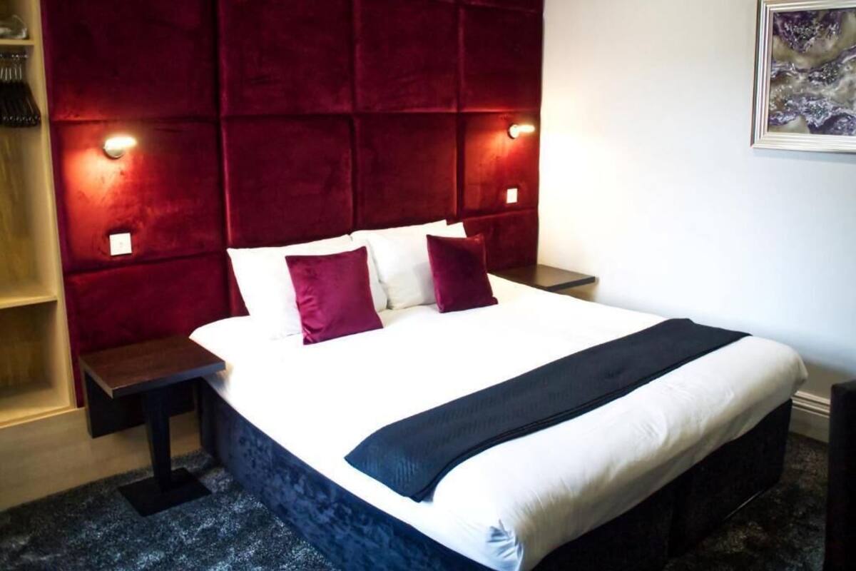 Une chambre d’hôtel avec un grand lit, deux tables de chevet, une tête de lit recouverte de tissu rouge et un tableau au mur.