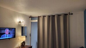 Un salón con una televisión de pantalla plana montada en la pared, una mesita de madera oscura con una lámpara y una cortina gris que cubre una puerta.