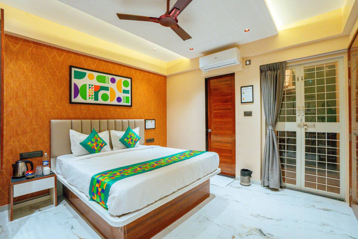 Habitación de hotel con cama, ventilador de techo, aire acondicionado en la pared, puerta con vistas y una mesita con tetera y tazas.