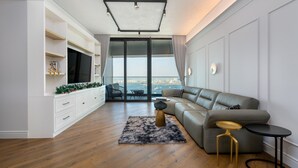 A modern living room with a large sofa, a flat-screen TV, a dining area with a table and chairs, and a view of the city through large windows.