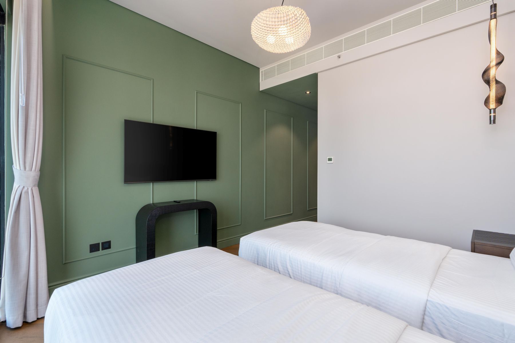 A hotel room with two beds, a flat-screen TV mounted on the wall, a modern pendant light, and a small table.