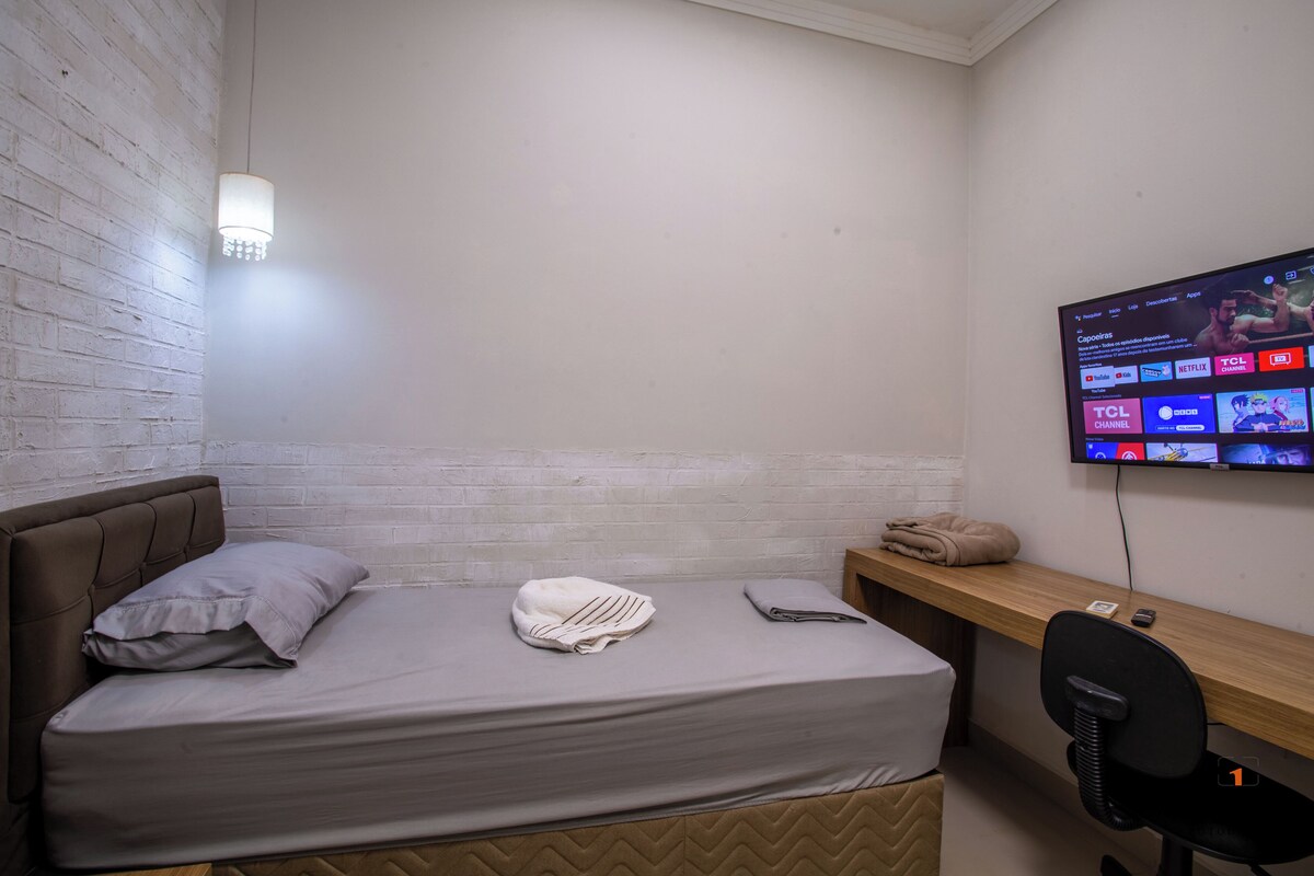 Un letto singolo con testiera, un comodino, una sedia e una TV a muro che mostra diversi canali.