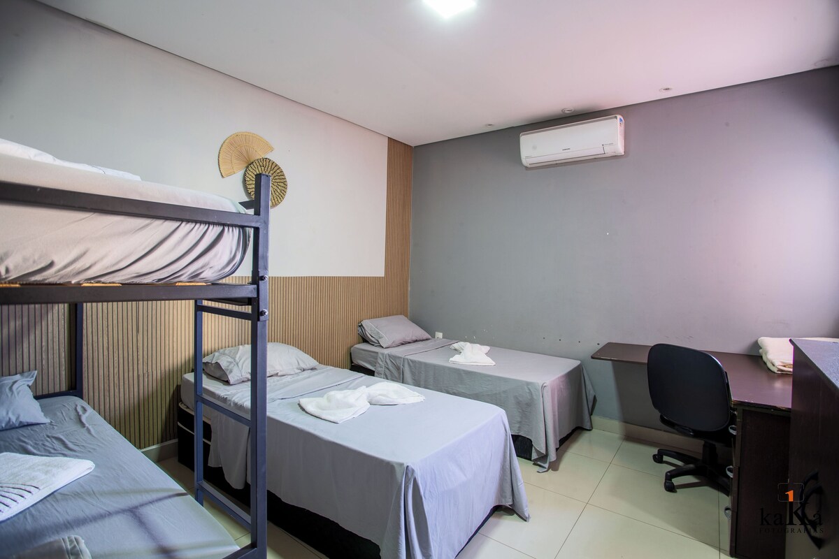 A dormitory room with bunk beds, two single beds, a desk, a chair, an air conditioner, and a wall fan.