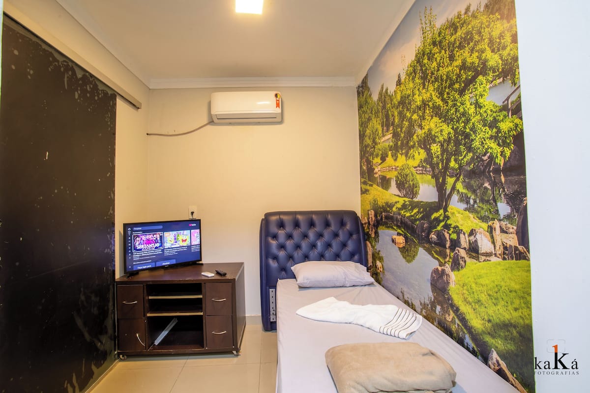 A bedroom with a bed, a TV on a dresser, an air conditioner, and a mural of a landscape.