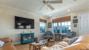 A living room with a large flat-screen TV, a ceiling fan, and a bookshelf. - Sand Digs! Adorable Gulf Beachside, 2 Bedroom, 2 Bath, Pet Friendly! (Dauphin Island)