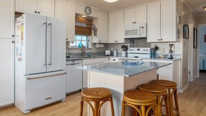 A modern kitchen with white cabinets, a stainless steel refrigerator, a microwave, and a large island with stools. - Sand Digs! Adorable Gulf Beachside, 2 Bedroom, 2 Bath, Pet Friendly! (Dauphin Island)