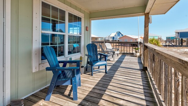 A wooden deck with two blue Adirondack chairs, a wooden railing, and a view of beach houses in the background. - Sand Digs! Adorable Gulf Beachside, 2 Bedroom, 2 Bath, Pet Friendly! (Dauphin Island)
