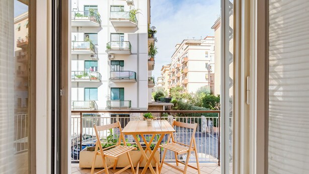 A balcony with a wooden table and chairs, a potted plant, and a view of neighboring buildings. - Cozy 2-Bedroom Flat with Balcony (Rome)