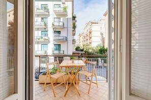 Un balcon avec une table et des chaises en bois, un pot de fleurs et une vue sur les bâtiments voisins.