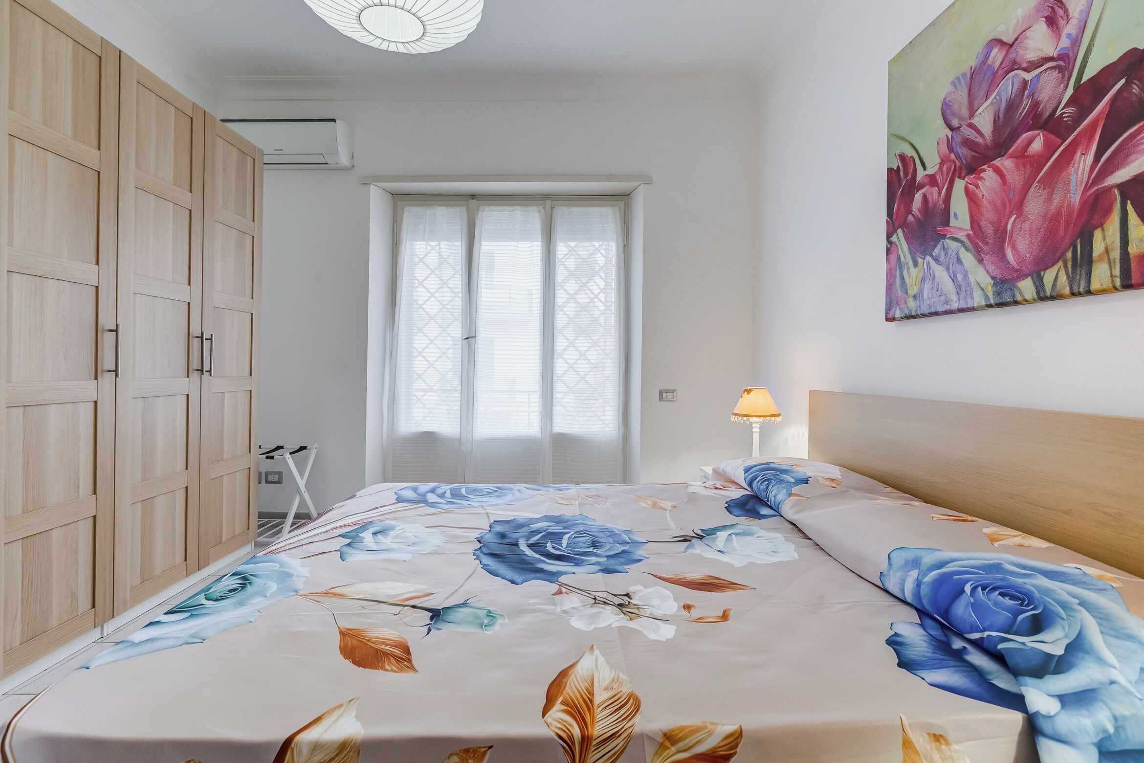 Un dormitorio con cama, armario de madera, ventana y un cuadro en la pared.