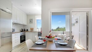 A modern kitchen with white cabinets and stainless steel appliances, a dining area with a table set for two, and a large window offering a view of the outdoors. - Fabulous apartment in beautiful Quinta do Paraiso (Carvoeiro)