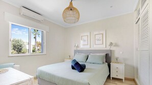 A bedroom with a bed, bedside tables, a desk, and a window with a view of palm trees. - Fabulous apartment in beautiful Quinta do Paraiso (Carvoeiro)