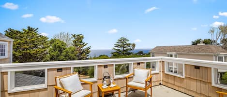 Eine Holzterrasse mit zwei weißen Sitzgelegenheiten und einem kleinen Tisch, mit Blick auf eine malerische Aussicht mit Bäumen und dem Meer.