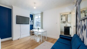 A compact living space with a blue sofa, a dining area with a table and chairs, a TV mounted on the wall, and a kitchen visible through an open door. - 3br-7guests-freeparking-pets-towncentre (Stoke-on-Trent)