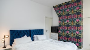 A bedroom with a blue tufted headboard, white bedding, a bedside lamp, a TV, and a floral-patterned curtain. - 3br-7guests-freeparking-pets-towncentre (Stoke-on-Trent)