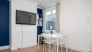 A room with a TV mounted on the wall, a window with curtains, a white table with chairs, and a small plant on the windowsill. - 3br-7guests-freeparking-pets-towncentre (Stoke-on-Trent)