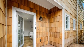 Une façade en bois avec une porte blanche, un petit espace extérieur aménagé pour s’asseoir et une fenêtre bien visible.