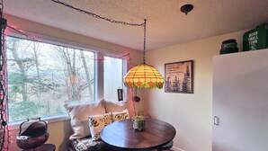 A cozy dining area with a round table, two chairs, a bench with cushions, a hanging lamp, and a window with a view of trees. - Charming 1 bedroom condo-walking distance to slopes (Sugar Mountain)