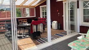 Et overdækket udendørsområde med træterrasse, et bord med stole og en loungeseng.