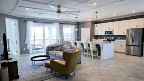 Amplia sala de estar con sofá, sillón y mesa de comedor. Se ve una cocina con electrodomésticos modernos y un balcón.