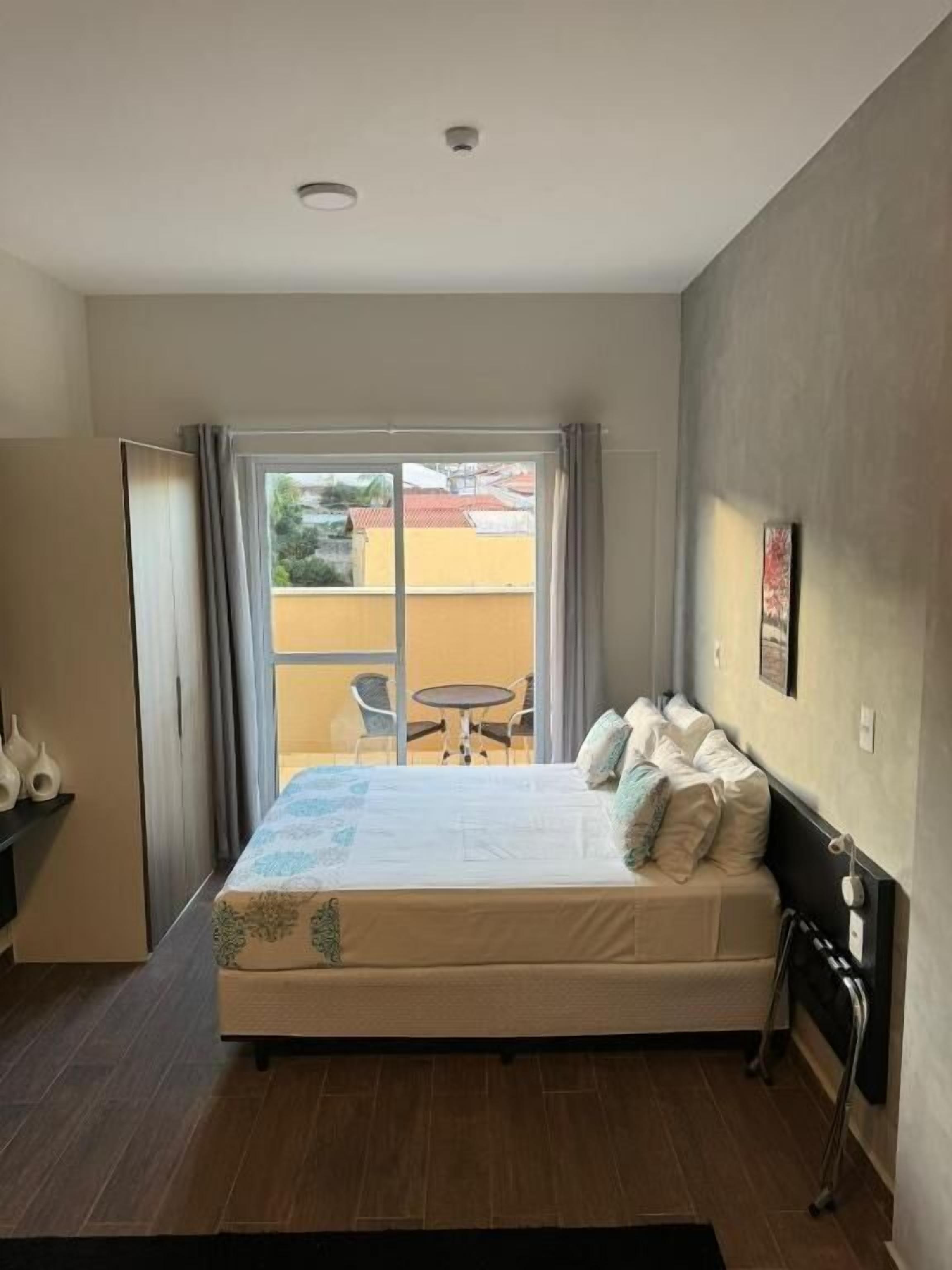 A hotel room with a bed, a small table, and a view of the outside.