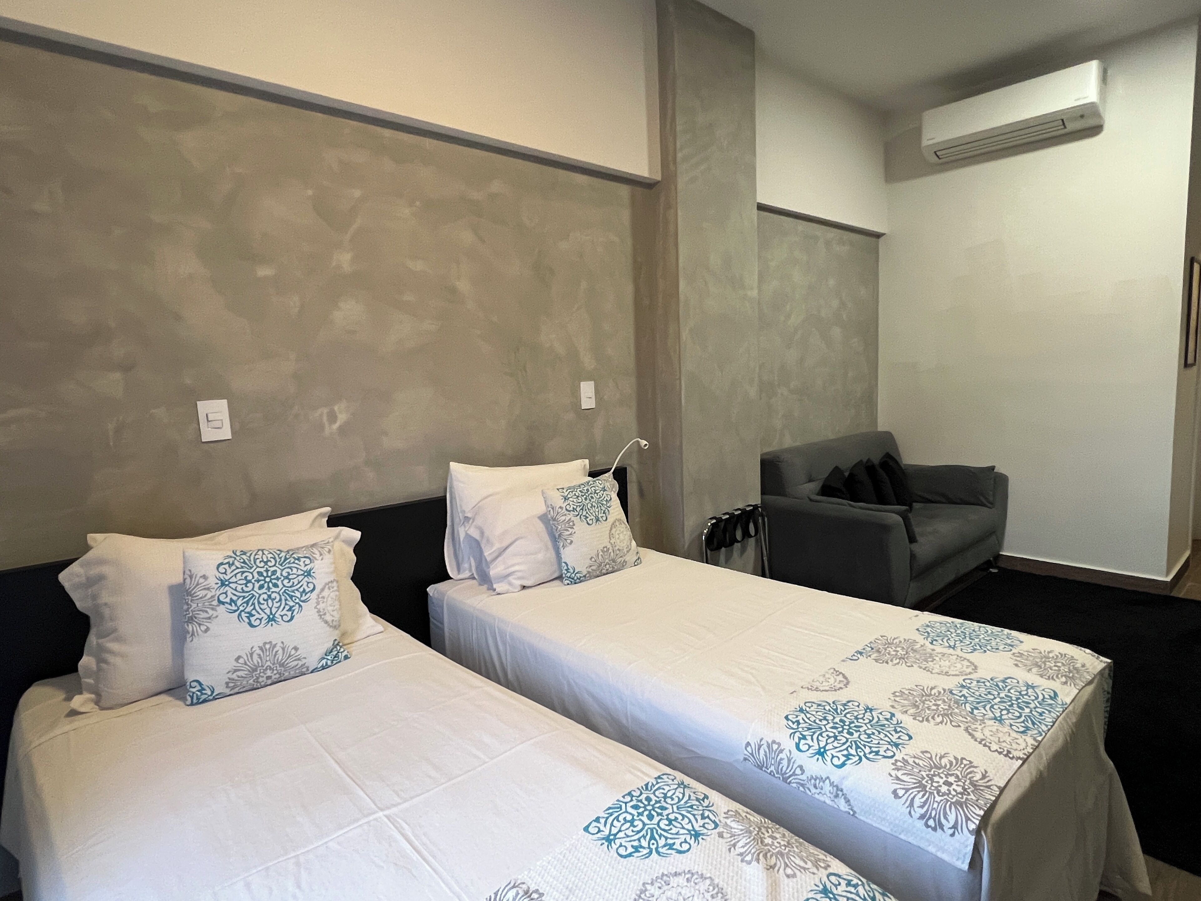 Una habitación de hotel con dos camas, un sofá gris y un equipo de aire acondicionado.