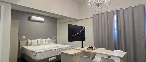 A modern hotel room with a bed, desk, two chairs, a TV, and a chandelier.