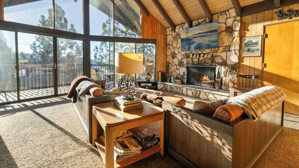A cozy living room with a stone fireplace, large windows, and a wooden ceiling.