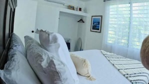 A hotel room with a bed, pillows, a ceiling fan, an air conditioning unit, a framed picture on the wall, and a window with curtains.