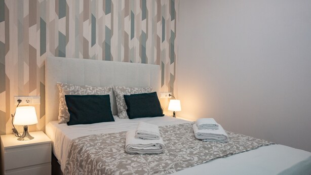 A neatly made bed with a patterned bedspread, two pillows, and two folded towels on a nightstand with a lamp.