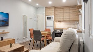 A modern living room with a sofa, a dining table with chairs, a TV, and a kitchen area with cabinets and appliances.
