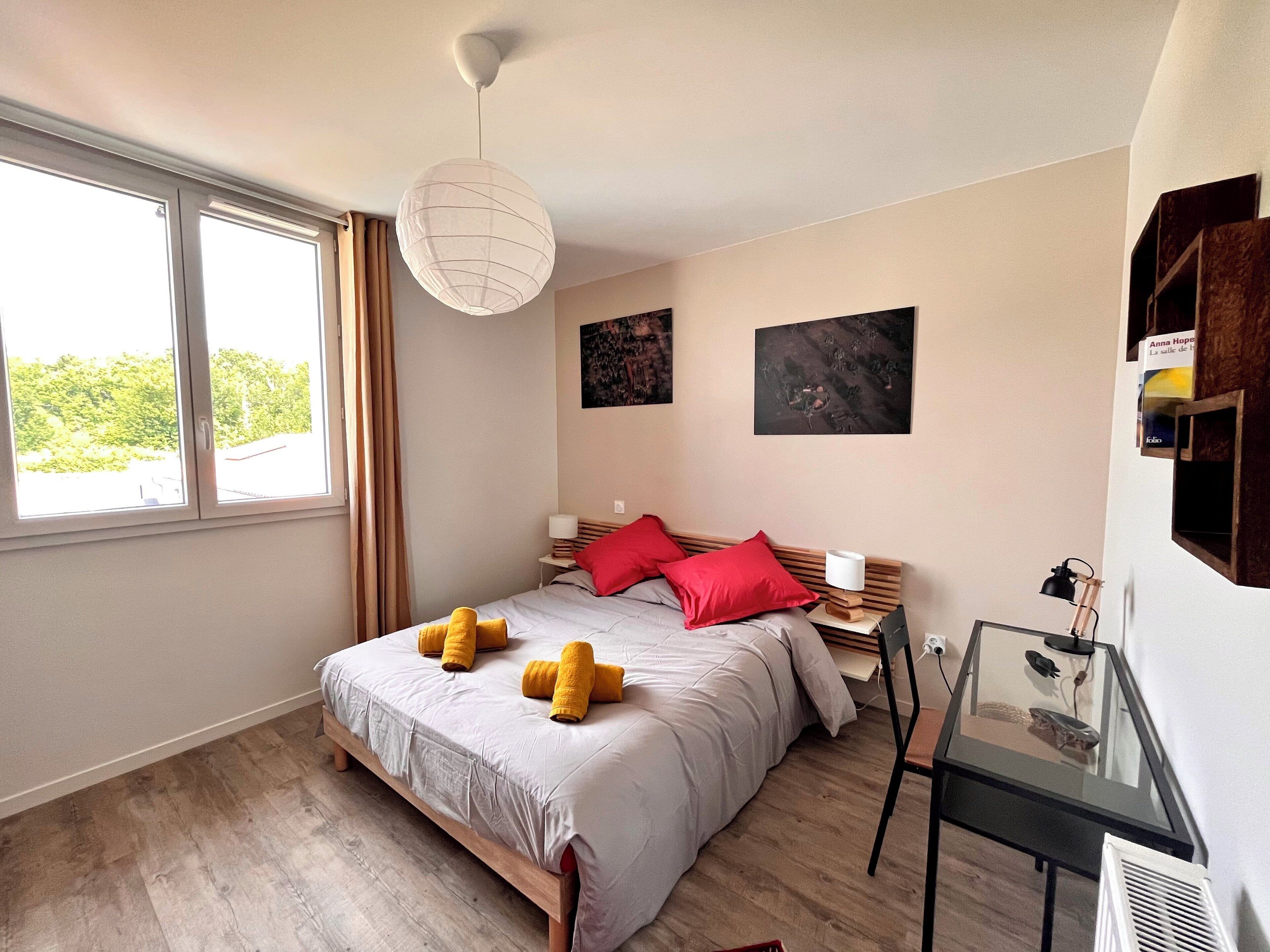 Ein Schlafzimmer mit einem großen Bett, einem Nachttisch mit Lampe, einem kleinen Tisch mit einem Dekorationsgegenstand und einem Fenster mit Vorhängen.