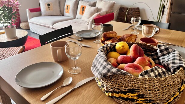 Une table à manger dressée avec des assiettes, des couverts, des verres et un panier rempli de fruits et de viennoiseries.