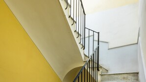 Een trap met marmeren treden en zwarte metalen leuningen, die uitkomt op een plateau met een witte muur en aan weerszijden een gele muur.