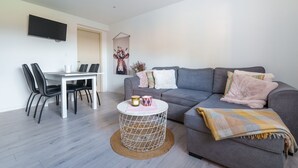 Una sala de estar moderna con un sofá gris, una mesa de comedor blanca con sillas, un televisor montado en la pared y una planta decorativa.