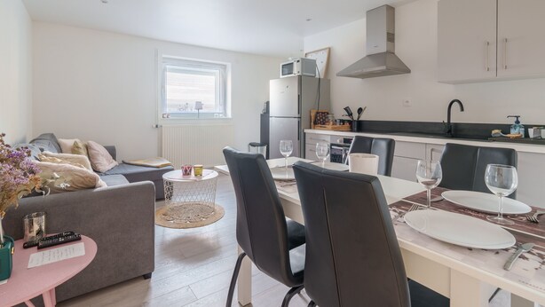 Una sala de estar moderna con un sofá gris, una mesa rosa y un comedor con una mesa blanca para dos personas.