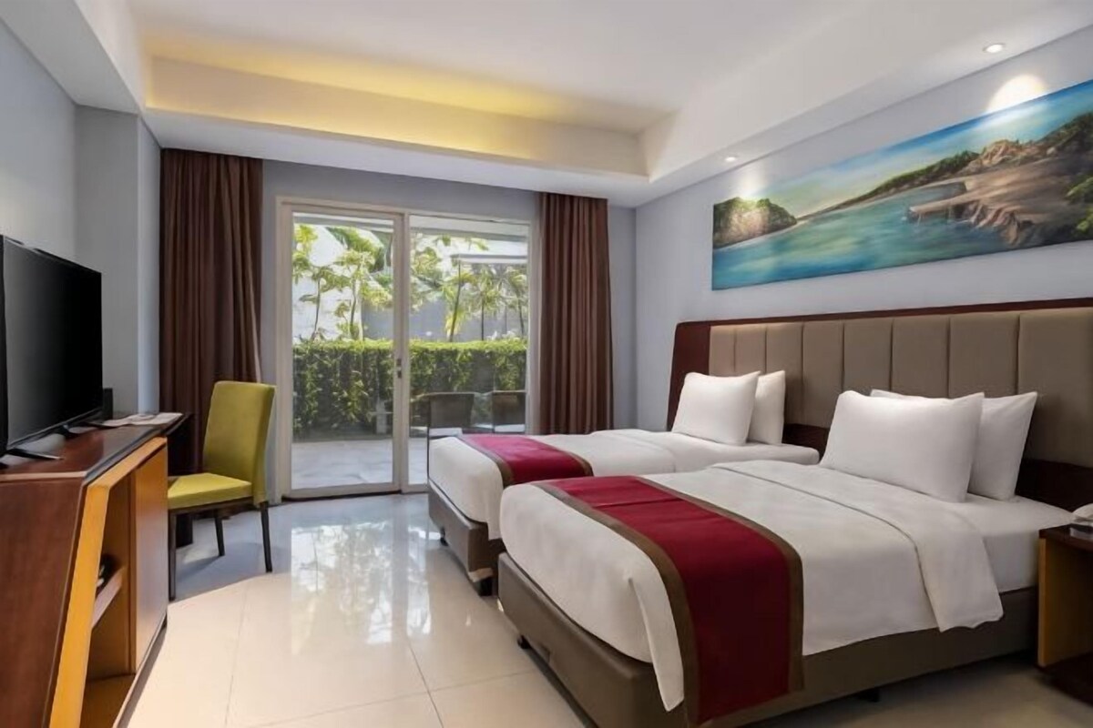 A hotel room with two beds, a TV, a desk, and a balcony with a view of greenery.