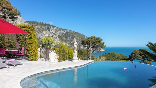 Ein Pool mit gewölbter Kante, umgeben von einer Terrasse mit Liegestühlen und einem roten Sonnenschirm.
