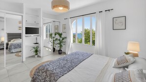 Ein modernes Schlafzimmer mit einem großen Bett, einem Fernseher, Topfpflanzen und Meerblick.