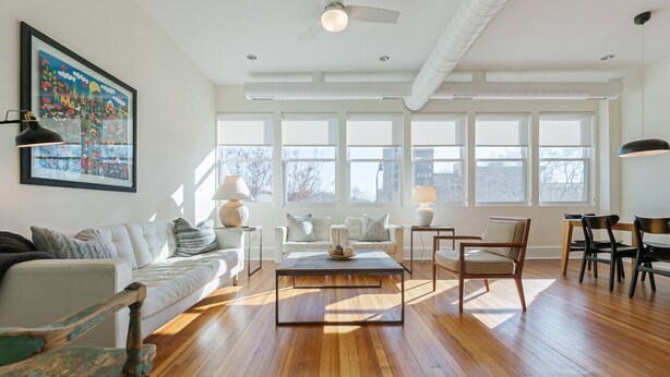 A spacious living room with hardwood floors, a white sofa, a coffee table, and a dining area with chairs. Large windows allow natural light to fill the space. - The Gazette Condo 200 at ATB (Asheville)