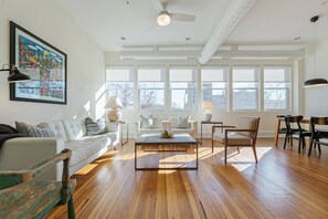 A spacious living room with hardwood floors, a white sofa, a coffee table, and a dining area with chairs. Large windows allow natural light to fill the space. - The Gazette Condo 200 at ATB (Asheville)
