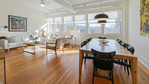 A spacious living room with a wooden dining table, a white sofa, and a large window with a view of the outdoors. - The Gazette Condo 200 at ATB (Asheville)