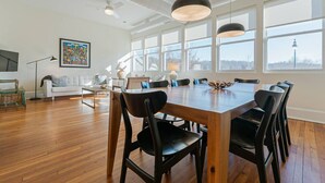 A spacious room with a large wooden dining table, black chairs, and pendant lights. There is a white sofa and a floor lamp in the background. - The Gazette Condo 200 at ATB (Asheville)