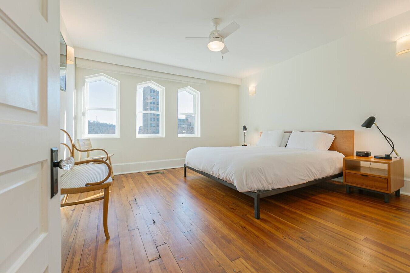 Un dormitorio con cama, mesitas de noche, una silla y ventanas amplias.
