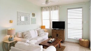 A living room with a white sofa, a wooden coffee table, a television, and a window with blinds.