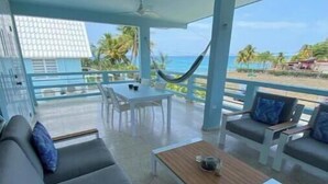 A covered outdoor seating area with a sofa, chairs, and a table, overlooking a beach with palm trees and a clear sky.