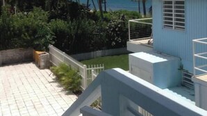 A small, single-story building with a tiled roof, surrounded by lush greenery and palm trees, overlooking a body of water.